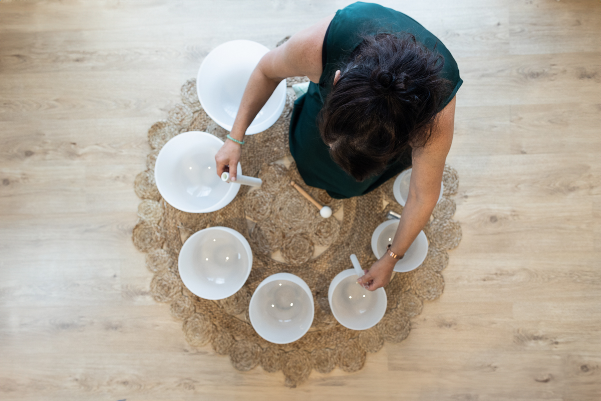 Overhead view of Vanessa Kenny from Soulstar Sound Healing seated on a woven mat, surrounded by white crystal singing bowls arranged in a circle. She wears a dark green dress and uses mallets in both hands to play the bowls in a calm, wooden-floored studio. The image captures the essence of Sound Healing and Reiki in NZ, showcasing a peaceful and meditative practice.