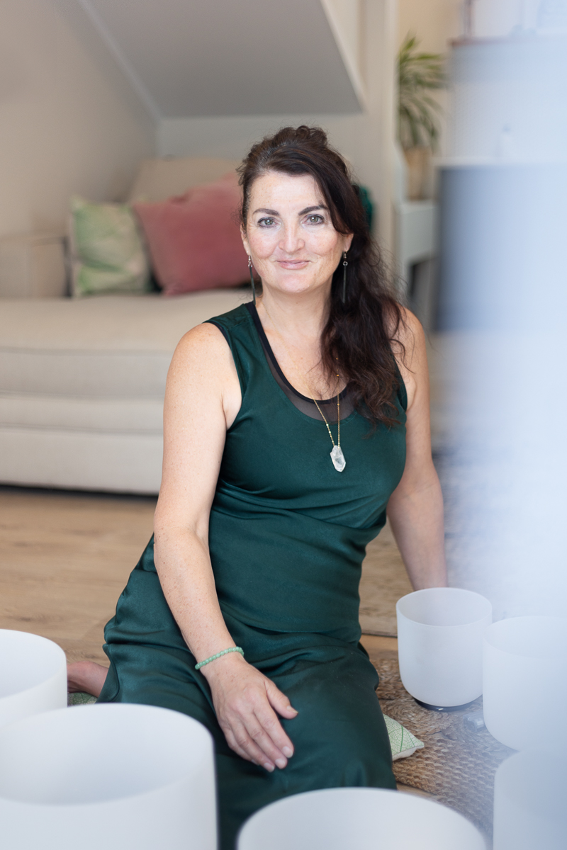 Vanessa Kenny of Soulstar Sound Healing sits peacefully on a woven mat surrounded by white crystal singing bowls. She wears a dark green dress and a clear quartz pendant, with a soft smile in a calming indoor space. This portrait captures the serene energy of her Sound Healing and Reiki practice in NZ.